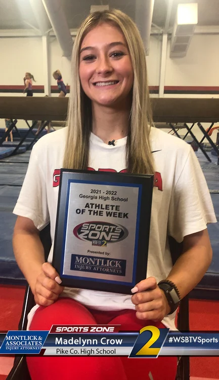 Teen gymnast smiles holding Athlete of the Week award from a local sports program at her high school gymnasium.