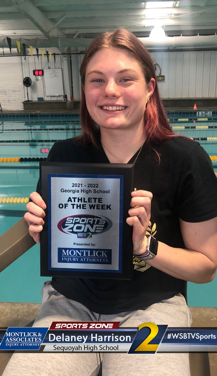 Smiling high school athlete of the week award winner poses in a swimming pool area holding her plaque.
