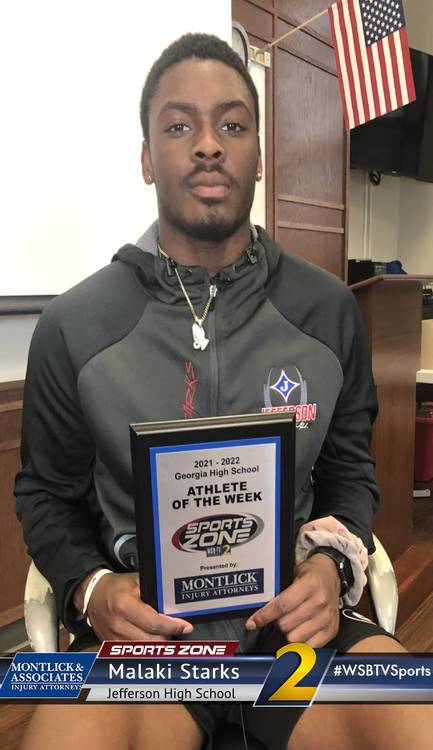 African American male student athlete of the week holding award plaque in classroom with American flag