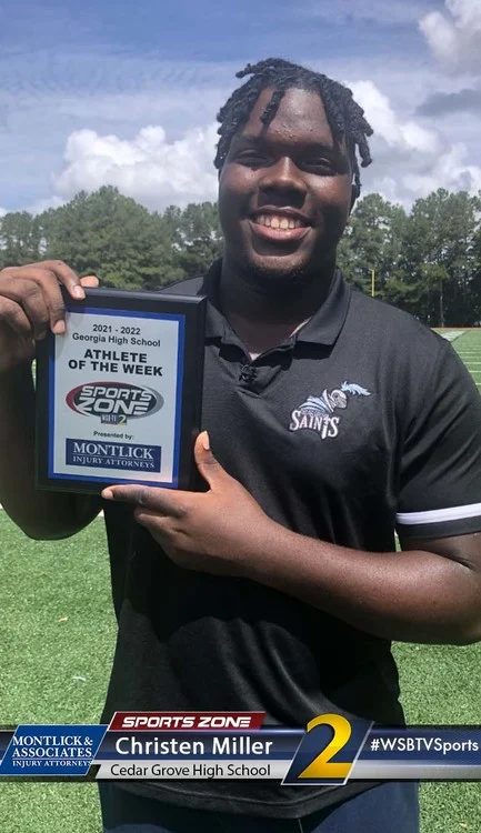 Black male athlete smiling and holding an "Athlete of the Week" plaque on a football field.