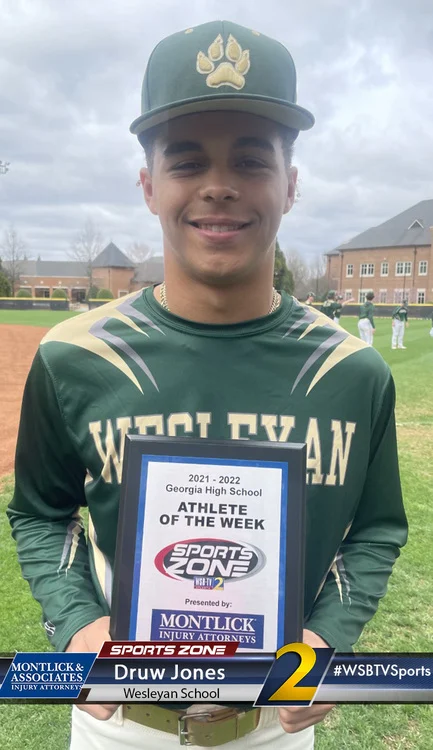 Young male athlete holding Athlete of the Week award on baseball field, smiling at camera.