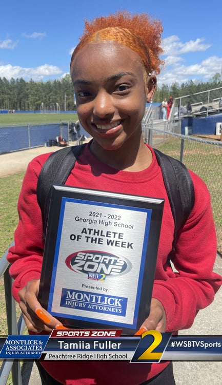Smiling young female high school athlete holding Sports Zone Athlete of the Week award on sports field.