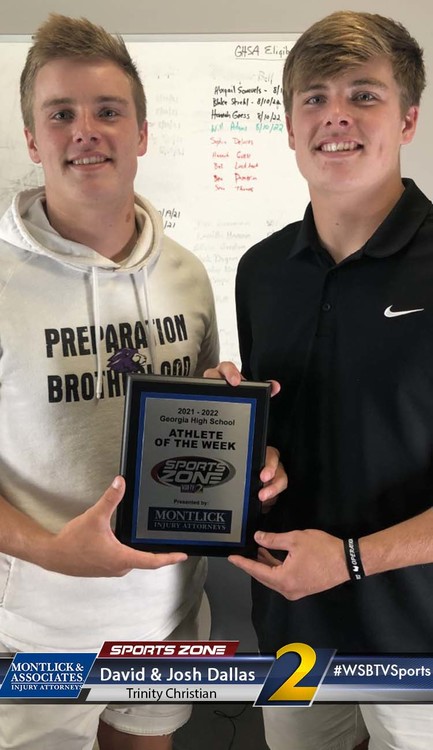 Two smiling male student athletes holding an "Athlete of the Week" sports award plaque, dressed casually.