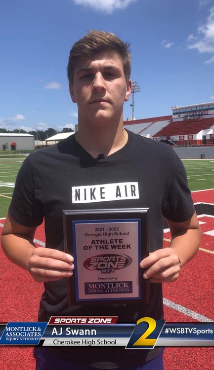 Young male athlete holding an award plaque while standing on a red track field under a blue sky.