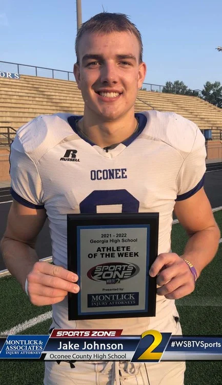 Smiling high school football player holding Sports Zone Athlete of the Week award plaque on field