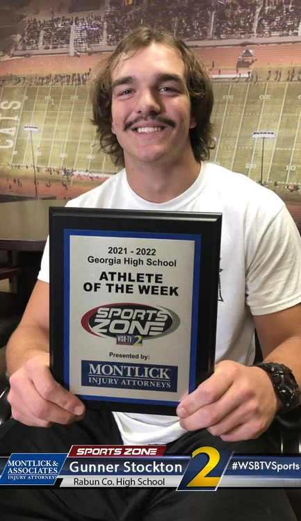 Smiling male high school athlete holds Sports Zone Athlete of the Week award in stadium bleachers.