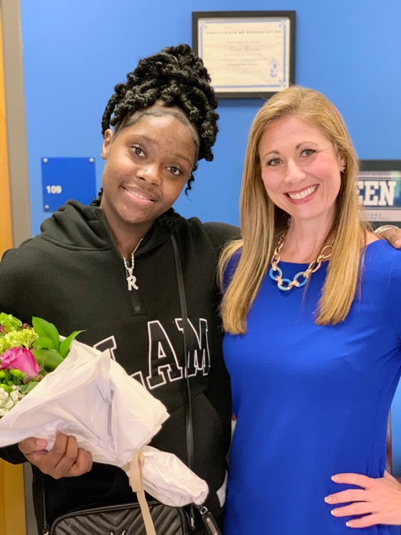 Two smiling women, one in a blue dress and one in a "LAW" hoodie, standing together holding flowers in an office setting.