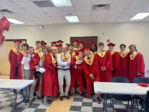 Group of high school graduates in red cap and gowns pose together in classroom, smiling at camera
