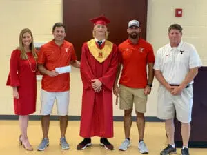 Red and white dressed school personnel flank graduate in cap and gown holding diploma, smiling proudly.