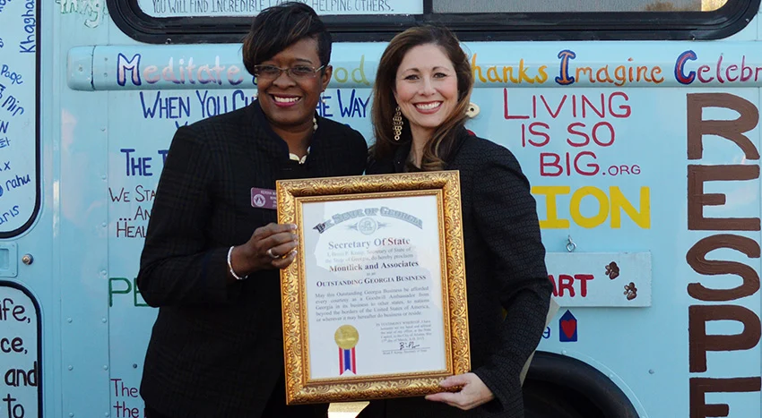 Two smiling women hold an award certificate in front of an inspirational mural with uplifting phrases.