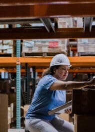 Woman worker wearing hard hat lifting box in warehouse storage area (SEO alt text, 69 characters)