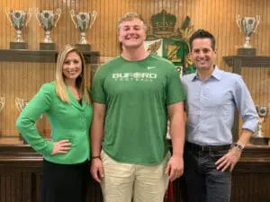 Smiling athlete in Buford Football shirt with coaches, trophies behind, for sports injury attorney site.