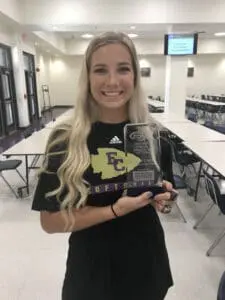 Smiling blonde female athlete holding softball trophy in school cafeteria, wearing East Central uniform