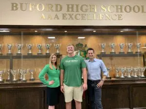 Alt text: Student athlete with parents in front of Buford High School trophy case displaying AAA excellence