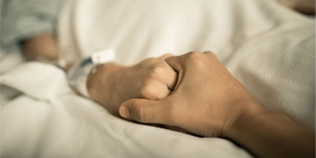 Close-up of hands clasped together, resting on a bed, showing support and comfort during difficult times.