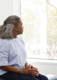 Elderly woman sitting near window, looking thoughtful. Informational medical posters in background.