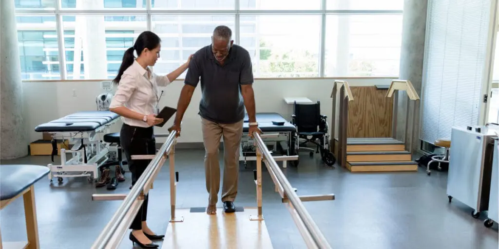 Injury attorney assisting patient with parallel bars during physical therapy session in clinic.