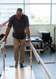 Injury attorney assisting patient with parallel bars during physical therapy session in clinic.