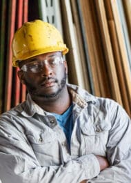 Construction worker in hard hat looking serious, arms crossed in warehouse with lumber