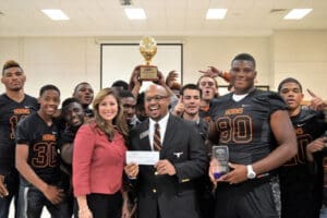 High school football team presents award to smiling man and woman in an office setting.