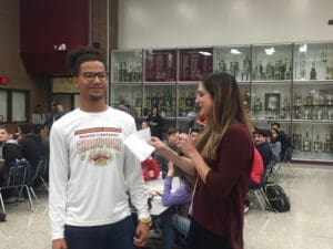 High school students smiling while examining championship rings in a school trophy case. The image shows an excited male student wearing a sports sweatshirt and a smiling female student looking at large athletic championship rings.