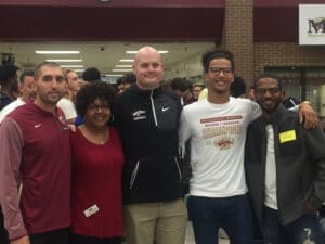 Group photo of diverse smiling people, one wearing a football championship t-shirt, at an indoor event