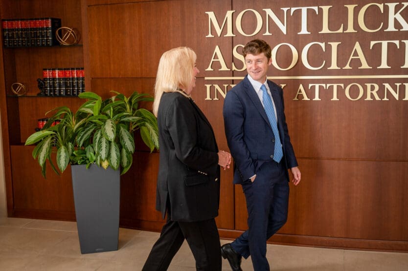 A smiling man in a suit shakes hands with a blonde woman in front of a sign reading "Montlick Associates Injury Attorneys".