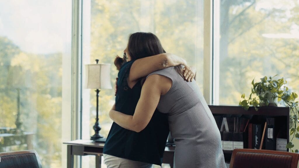 Woman hugging herself, looking out window, suggesting emotional distress or trauma after an injury.