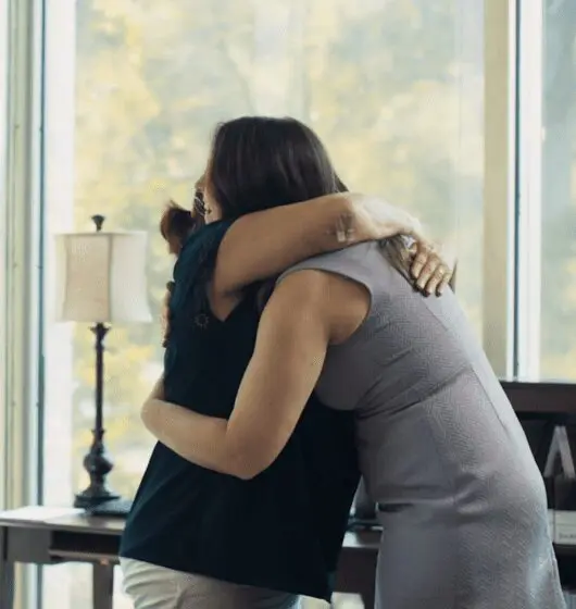 Woman hugging herself, looking out window, suggesting emotional distress or trauma after an injury.