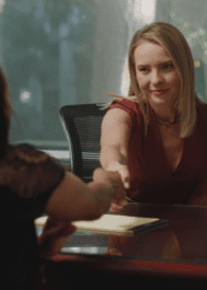 The image shows a young blonde woman smiling and shaking hands with someone across a desk in an office setting. The woman appears to be a professional, possibly a lawyer or consultant, meeting with a client or colleague.