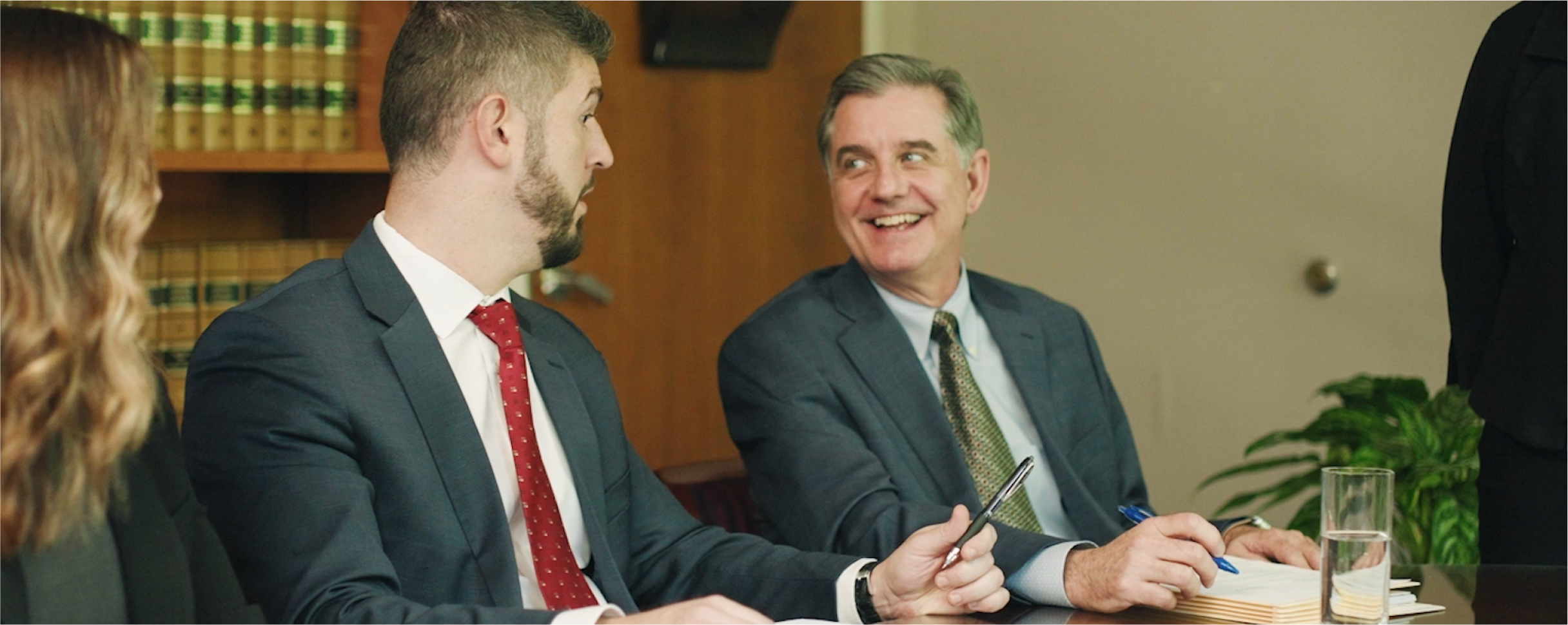 Two smiling businessmen in suits discussing at an office table, suggesting a legal consultation meeting.