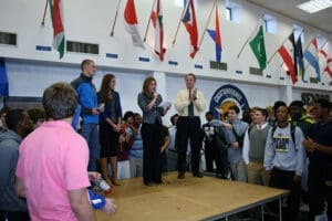 The image shows a group of students gathered in what appears to be a school gymnasium or auditorium, with various international flags hanging from the ceiling. Some adults, likely teachers or administrators, are standing amidst the students. The students seem to be focused on or interacting with the adults. Given the presence of the flags, this event may be some type of international or cultural gathering at the school. However, without more context it's difficult to determine the exact nature or purpose of the assembly.