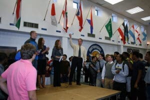 School assembly with international flags; students gather around adult speakers at podium