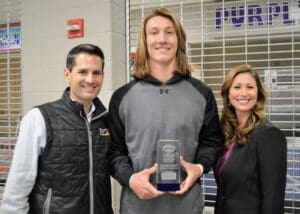 Injury attorney with tall smiling athlete holding award plaque, standing between two others at a sporting venue.