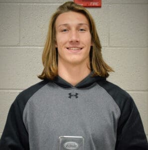 Young smiling male athlete in gray Under Armour hoodie standing in front of red fire alarm pull station.