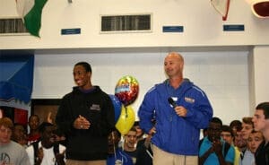 Two men stand in a gym, one holding balloons, in front of a crowd of young athletes. The image shows community involvement, likely as part of an outreach or mentorship program put on by the attorney's office to engage with local youth through athletics.