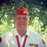 Middle-aged man in red fez smiling outside surrounded by green foliage, representing Shriners organization.