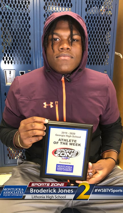 Smiling high school athlete of the week holding Sports Zone award in locker room, sponsored by injury attorneys.
