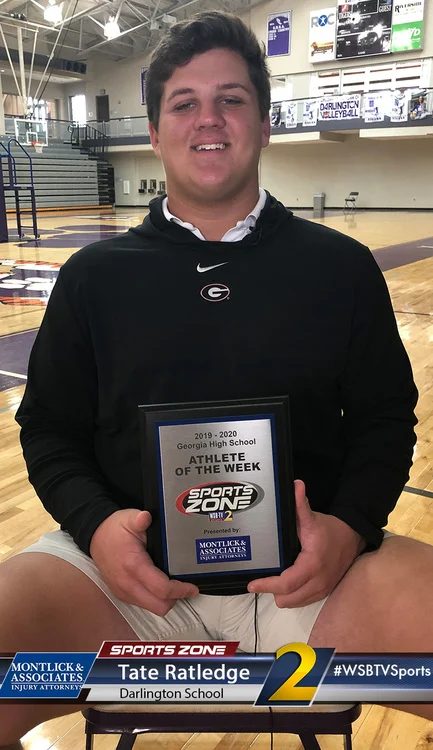Young male athlete holding Athlete of the Week award in high school gym, smiling at the camera.