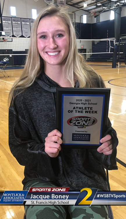 Smiling female athlete holding "Athlete of the Week" sports award in a high school gym setting.