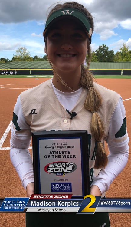 Smiling softball player in uniform holding "Athlete of the Week" award at home plate on baseball field.