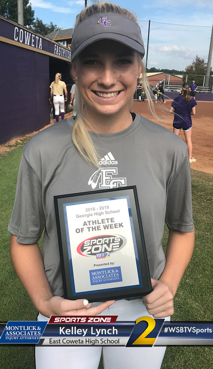 Smiling athletic high school girl holding Athlete of the Week award on sports field, wearing gray Adidas hat and shirt.