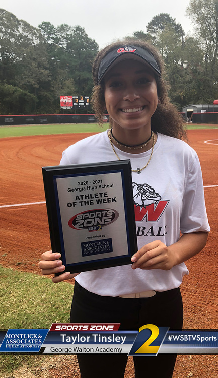 Smiling softball athlete holding Athlete of the Week award plaque on athletic field