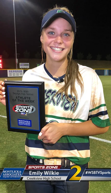 Athlete of the week Emily Wilkie holding Sports Zone award plaque on baseball field, smiling happily.