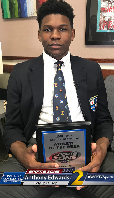 High school student athlete holds sports award plaque for Athlete of the Week, smiling in suit and tie.