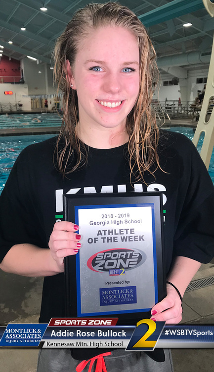 Young female swimmer smiling and holding Athlete of the Week award certificate at indoor pool.
