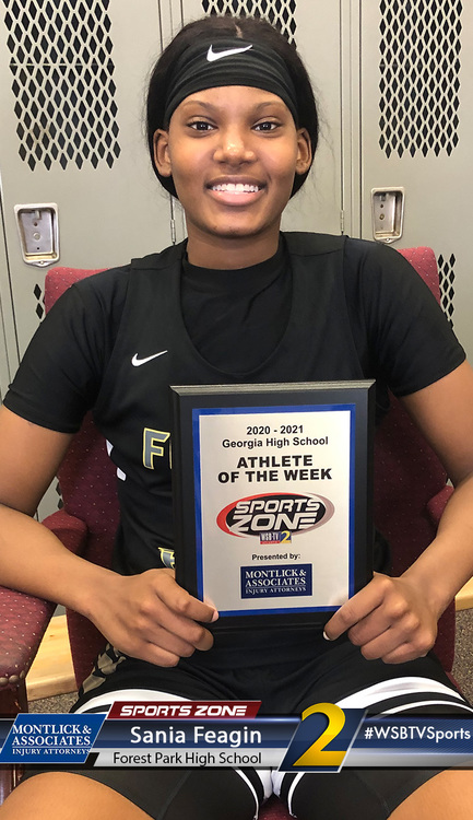 Athletic female high school student holding Athlete of the Week award smiles in locker room setting.