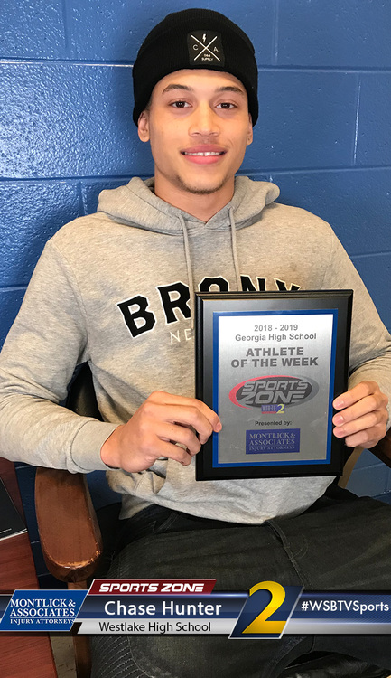 Smiling male high school athlete of the week holding award, wearing grey hoodie and black hat.