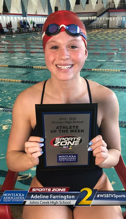 Smiling young swimmer holding "Athlete of the Week" award plaque at indoor pool, wearing swim cap and goggles.