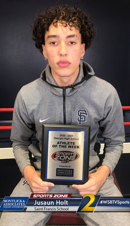 Young male athlete holding "Athlete of the Week" award, wearing a grey hooded sweatshirt, in a sports setting.